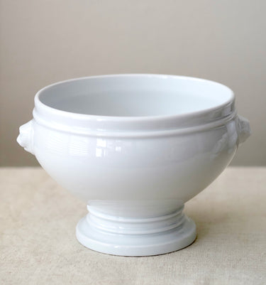 A glossy white ceramic pedestal bowl with side handles, set against a neutral beige background.
