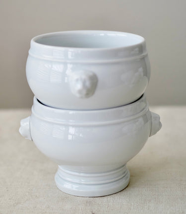 A pair of stacked white ceramic bowls with sculpted lion head handles and a pedestal base, set against a neutral beige background.