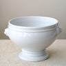 A glossy white ceramic pedestal bowl with side handles, set against a neutral beige background.