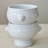 A pair of stacked white ceramic bowls with sculpted lion head handles and a pedestal base, set against a neutral beige background.