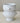 A pair of stacked white ceramic bowls with sculpted lion head handles and a pedestal base, set against a neutral beige background.