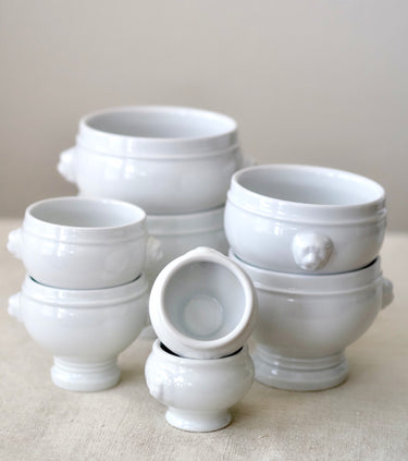A set of glossy white ceramic bowls with handles and footed bases, arranged in a stack on a light beige fabric surface against a neutral beige background.