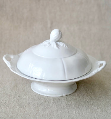 A white ceramic lidded serving dish with decorative side handles and a pedestal base, photographed against a neutral textured beige fabric background.
