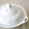 A white ceramic lidded serving dish with a decorative embossed floral knob on the lid and a side handle, photographed on a soft beige fabric background.