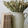 A stoneware confit pot filled with dried millet sits on a light fabric surface next to a stack of vintage French rag books in a deep brown color. 
