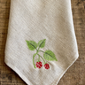 A cream-colored linen kitchen towel with embroidered green leaves and raspberry motif, laid flat on a rustic wooden surface.
