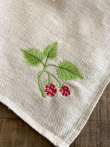 A close-up of a cream-colored linen fabric tea towel corner featuring an embroidered green leafy branch with a red raspberry motif, resting on a dark wooden surface.